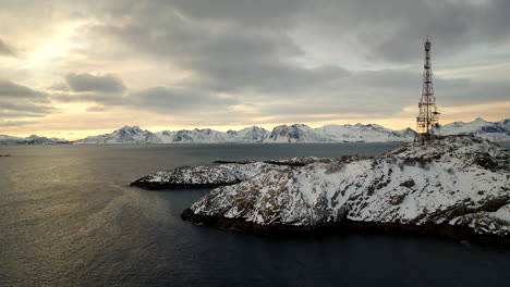 Telecommunication-tower-on-snowy-rock-island-in-Henningsvaer,-sunrise-aerial