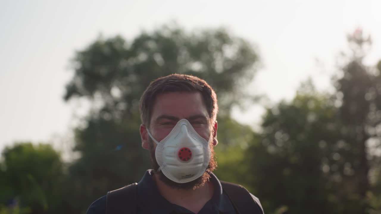 Primo piano di un uomo che irrora le colture con uno spruzzatore a zaino, indossando maschera e guanti sotto la luce dorata del sole che crea un bagliore, mentre esegue fumigazione e controllo dei parassiti all'aperto su piante a foglia.