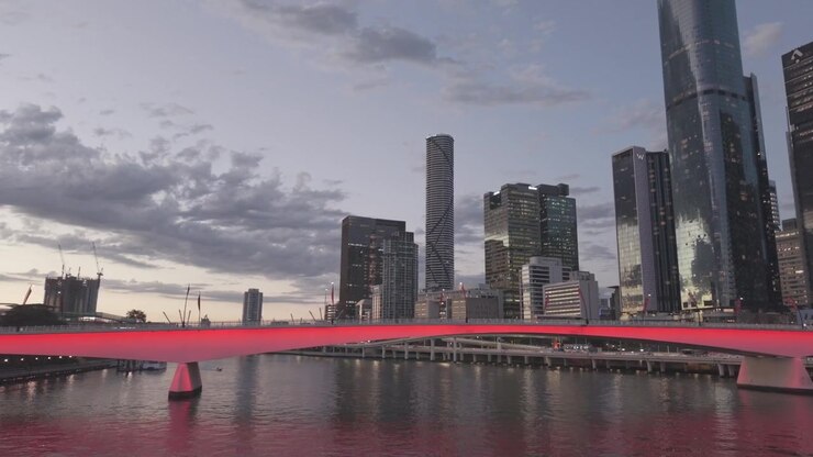 Uma vista aérea ao entardecer mostra a Ponte Victoria brilhando com luz vermelha ao longo da orla fluvial do CBD de Brisbane