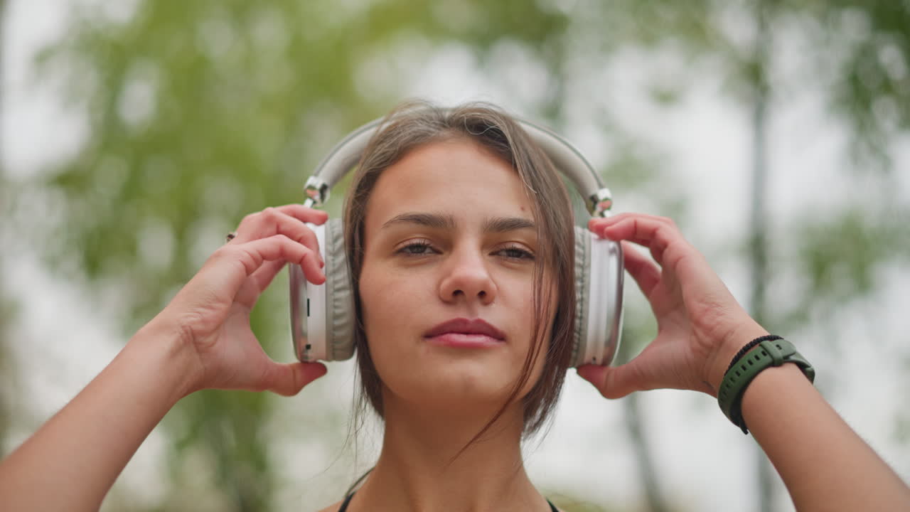Portrait of woman placing headphones on her head in outdoor environment with trees blurred in the background, she has a relaxed expression, preparing to listen to music while enjoying fresh air