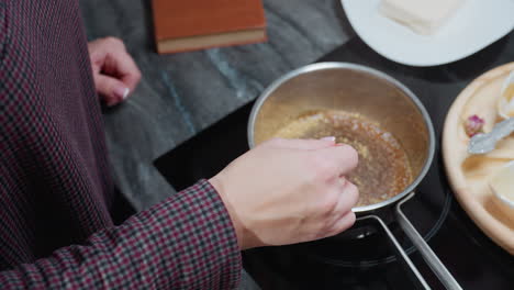 partial top-down view of person stirring soup in stainless steel pot using a stick on electric stove, with wooden tray, utensils, and ingredients arranged on dark marble countertop in modern kitchen