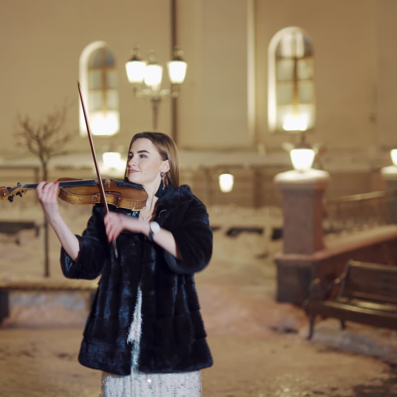 A beautiful girl is playing the violin in the centre of the city in the winter near the road on amid passing public transport. Blurred background.