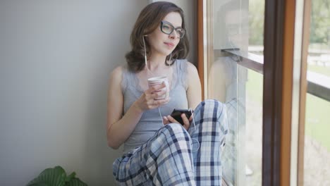 Young-woman-sitting-by-window-using-smartphone-with-earphones