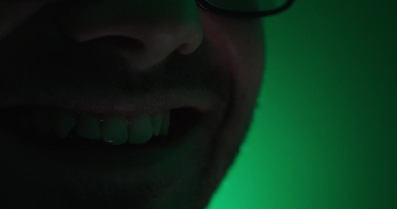 Close-up of a smiling mouth in dramatic green lighting, representing cyberbullying, online harassment, malicious behavior, and internet trolling.