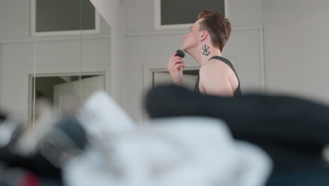 blurred grooming props on cushion bench include rope handcuff bag while tattooed neck boy applies makeup to jawline near open door and mirror reflection in bright minimalist studio interior