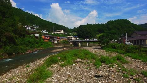 Serene-drone-shot-progresses-above-a-quaint-Japanese-village,-with-a-gentle-river-and-a-picturesque-bridge-crossing