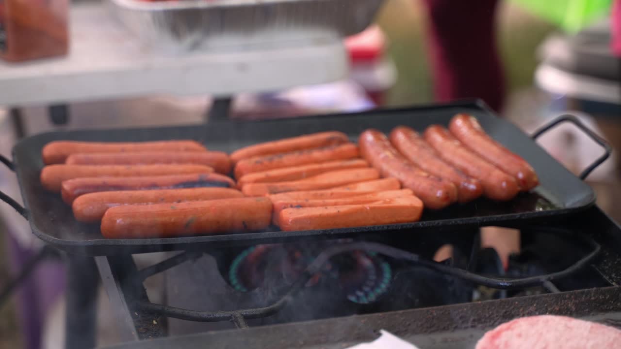 asar salchichas de perros calientes y salchichas vegetarianas en un plato caliente de hierro fundido sobre fuego de propano en la barbacoa al aire libre del campamento, mientras un hombre blanco caucásico coloca una hamburguesa en la parrilla