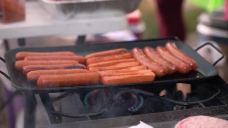 grilling hot dogs sausages and veggie wieners on cast iron hot plate over propane fire at outdoor campsite barbecue, as caucasian white man places hamburger on grill