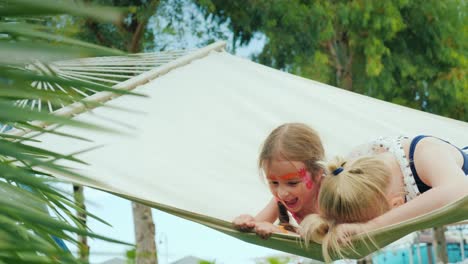 two cute funny sisters with painted faces rest on a hammock, have fun, laugh