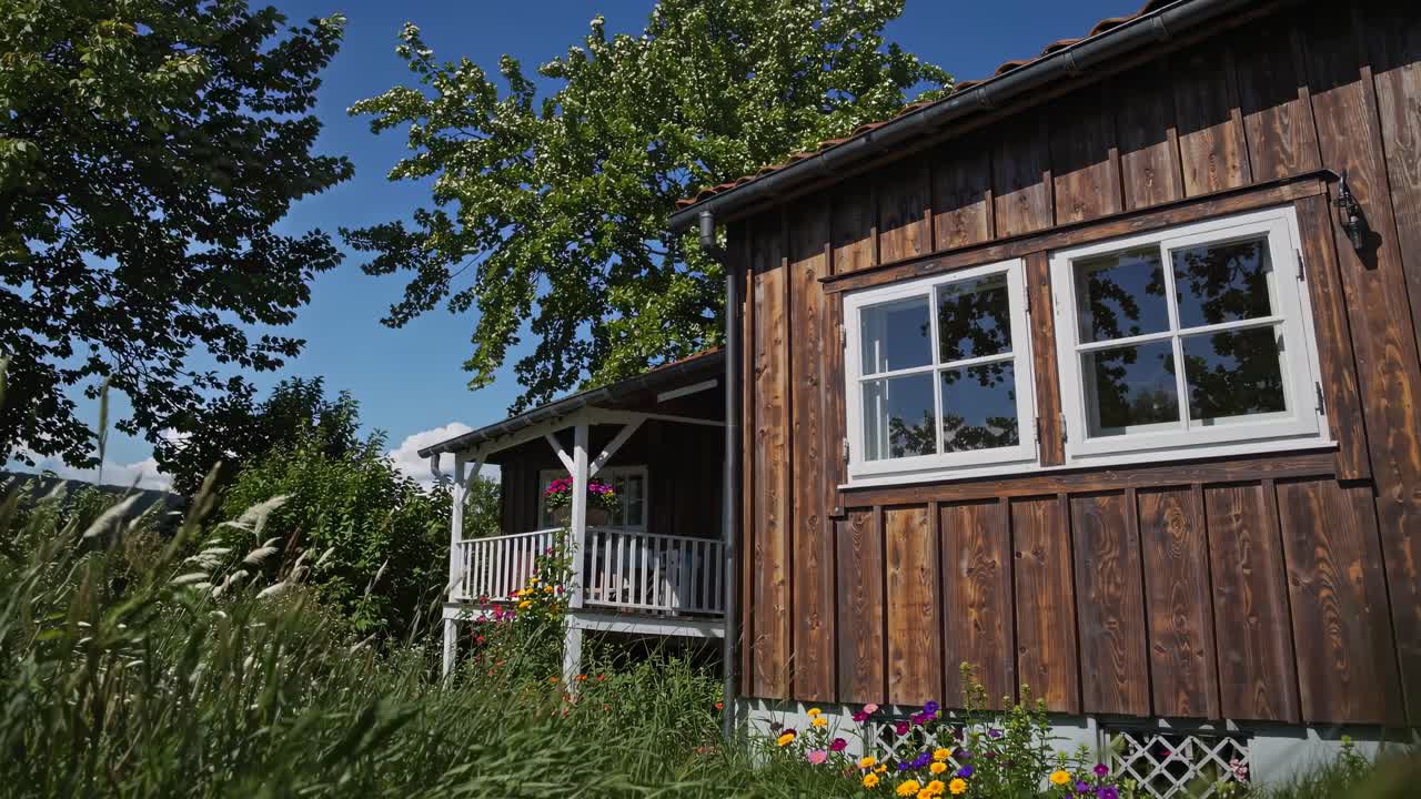 Riprese video di una casa rustica in legno con un portico, circondata da una vegetazione lussureggiante e fiori.