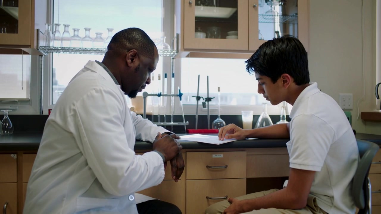 Estudiante señalando con polo blanco comenzando una revisión en la mesa de laboratorio, instructor con bata de laboratorio examinando un papel.