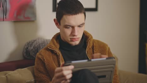 young caucasian man sitting on couch indoors and using tablet - medium shot