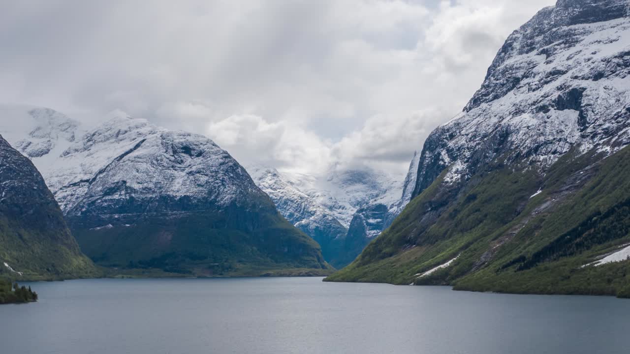 Thick, white clouds whirling above the calm waters of the Lovatnet lake, Norway. Ripples running on the water. Snowcovered mountains tower on the horizon. Forest is covering the shores of the lake