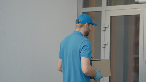 joven caucásico trabajador de entrega de hombre guapo con gorra azul caminando por la calle y llevando una caja de cartón mientras usa un teléfono inteligente buscando una ruta. mensajero masculino con paquetes tocando y enviando mensajes de texto por teléfono.