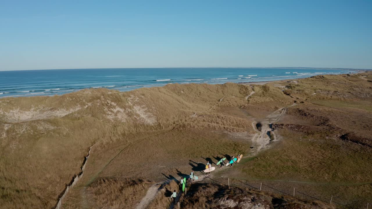 surfistas llevando tablas a lo largo de un sendero de arena en las dunas costeras, vista aérea