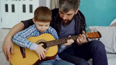 concentrated child is playing guitar with his father experienced guitarist, adjusting musical instrument and enjoying happy moments. music and family concept.