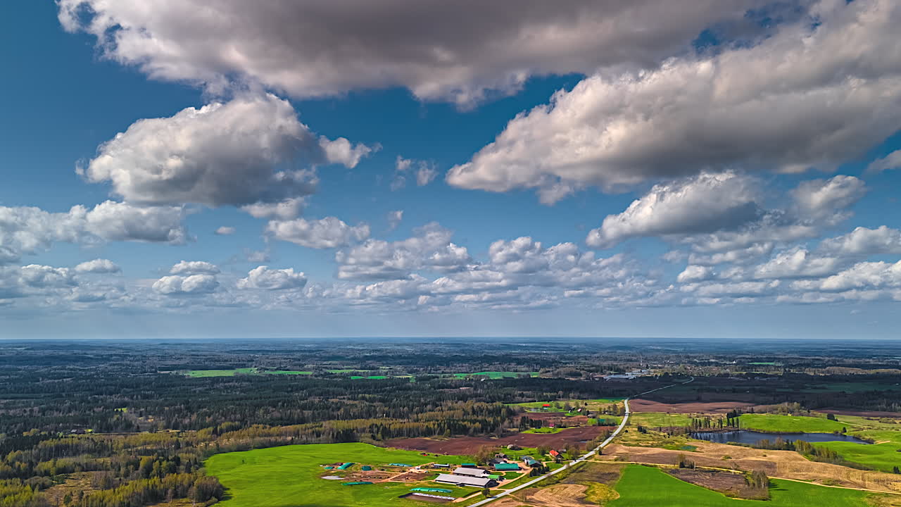 Rural, farmland and a highway passing through a European small town in a forested landscape under cloudscape casting shadows as they blow by in this aerial time lapse