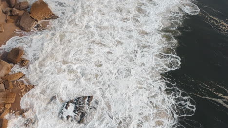 Aerial-view-of-waves-breaking-at-rocky-shoreline-in-Nazare,-Portugal