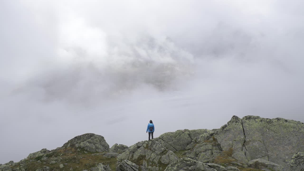 una excursionista solitaria se encuentra en el borde de un afloramiento rocoso con vistas a un gran glaciar alpino mientras la niebla y las nieblas se arremolinan a su alrededor