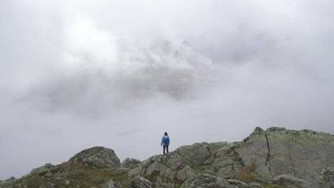 a lone female hiker stands on the edge of a rocky outcrop looking out over a large alpine glacier as the fog and mists swirl around her