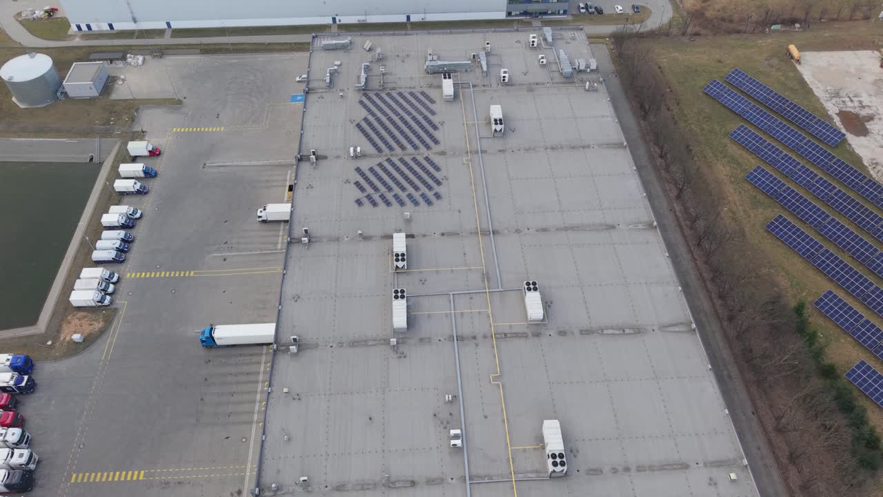 Aerial drone footage of a large logistics warehouse rooftop covered with solar panels alongside a ground mounted photovoltaic solar farm. Trucks visible at loading docks next to the industrial facilit