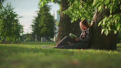 woman sitting outdoors with legs stretched, leaning against tree on grassy field, reading book under warm sunlight, tree leaves gently swaying in breeze, with background featuring greenery