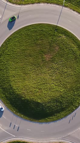 vertical drone static shot shows katlakalns roundabout with green central island, few car circulating, road markings, street lamps casting long shadows, spring fields around, slow motion