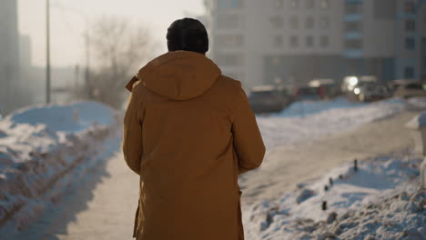 back view of young man wearing winter coat walking along snow covered sidewalk past parked cars under cold sky with footprints trailing behind frosted drifts and distant trees in morning light