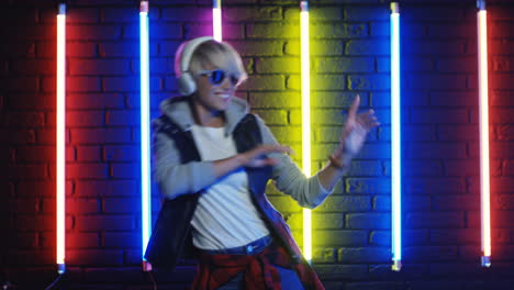 blonde woman in sunglasses and white headphones dancing in a room with colorful neon lamps on the wall