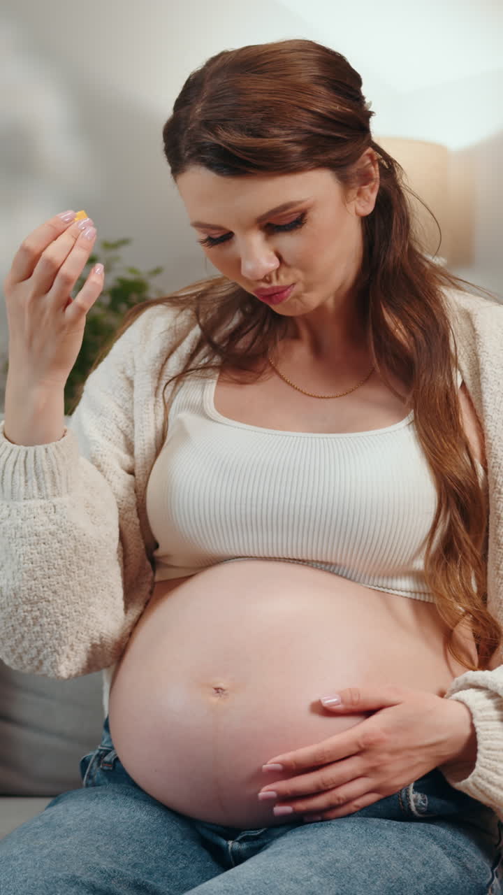 Pregnant woman holding belly and eating small yellow snack feeling content and relaxed at home