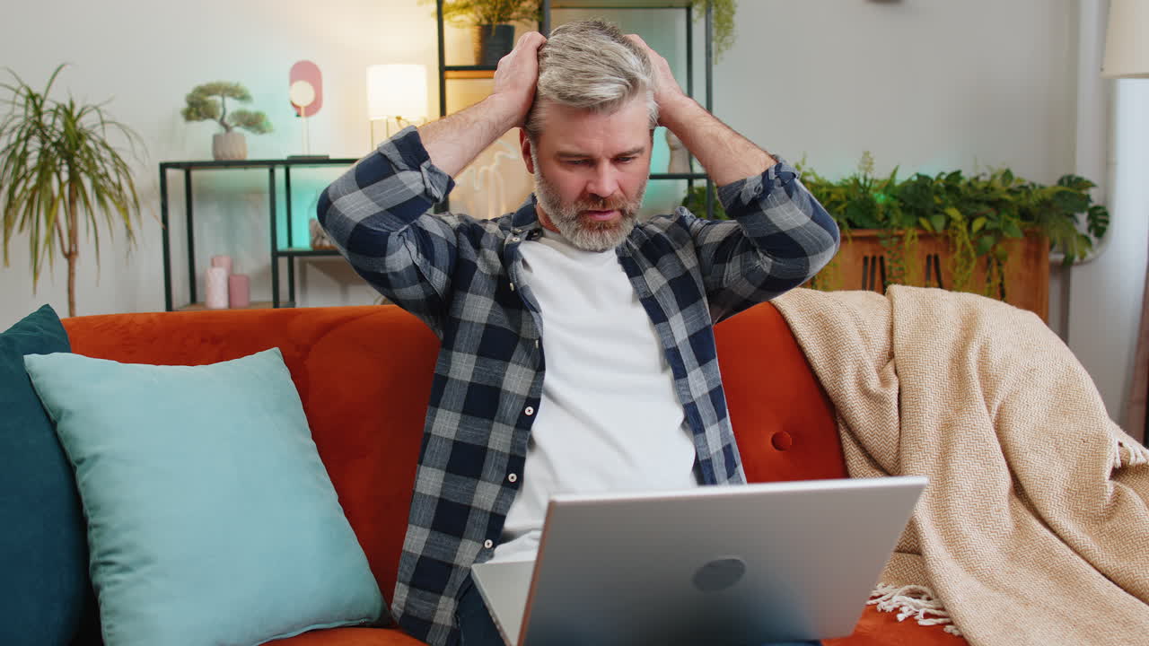 Upset senior man using laptop having anxiety stress reacting to loss bad news sitting on home sofa