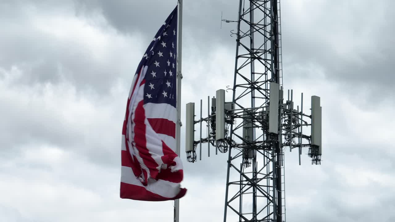 Winding american flag with red white stripes and stars beside transmission tower. Aerial close up. Cloudy day with Dark clouds at sky. Patriotic country of Trump.