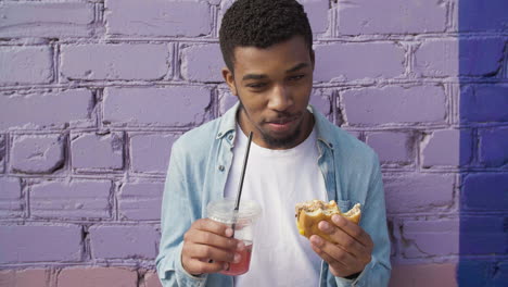 happy young man eating a delicious hamburger and holding a cold drink in plastic cup, leaning against a colorful brick wall in the city