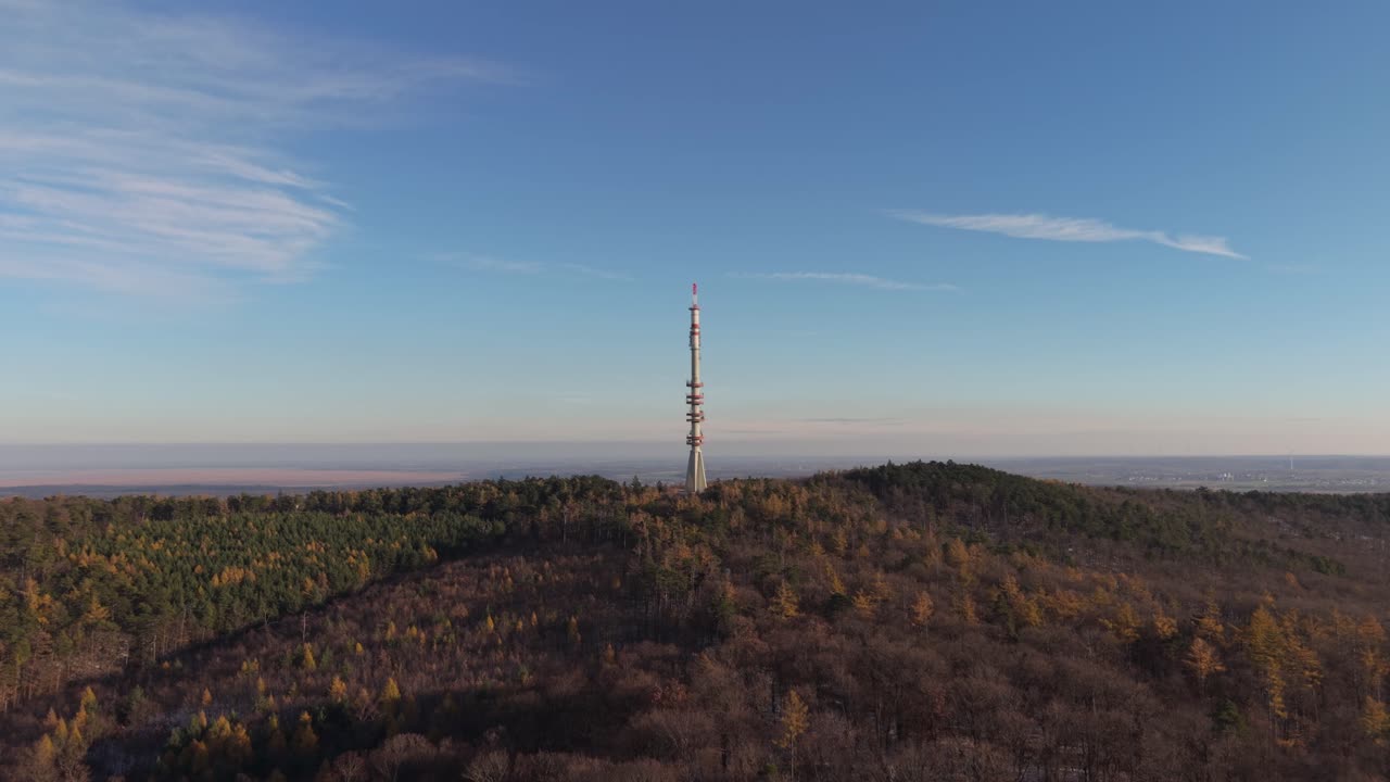 An aerial view of a tall telecommunication tower rising above a forested hillside. The scene captures the contrast between modern infrastructure and the natural landscape under a clear blue sky