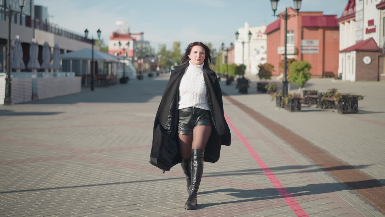 Confident woman strolling in black coat, white sweater, and black boots on paved urban street during sunny day, holding black handbag, surrounded by modern buildings
