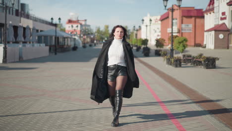 confident woman strolling in black coat, white sweater, and black boots on paved urban street during sunny day, holding black handbag, surrounded by modern buildings