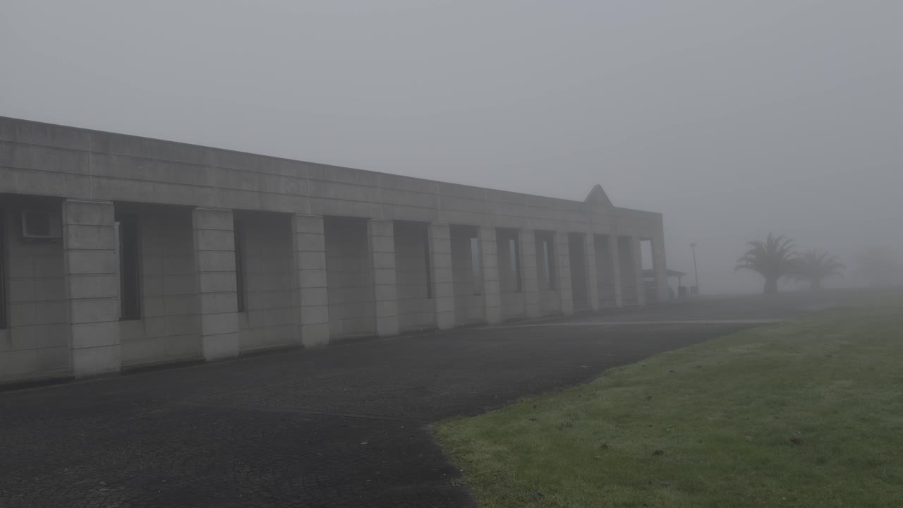 Spooky View Of A Modern Stone Building Covered In Fog Next To A Green Field