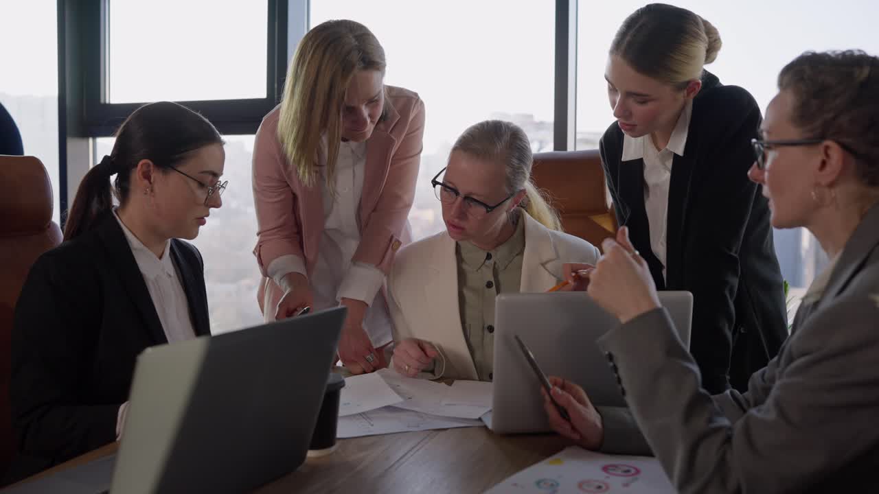 Confident middle-aged blonde girl with glasses in a white business suit communicates with her colleagues and listens to their ideas and suggestions about work in the office during a meeting with a wooden table in a modern office