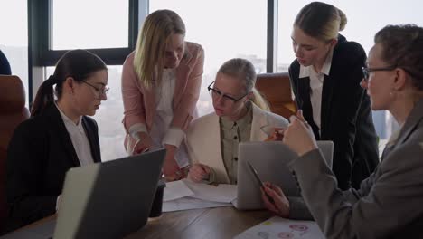 Confident-middle-aged-blonde-girl-with-glasses-in-a-white-business-suit-communicates-with-her-colleagues-and-listens-to-their-ideas-and-suggestions-about-work-in-the-office-during-a-meeting-with-a-wooden-table-in-a-modern-office