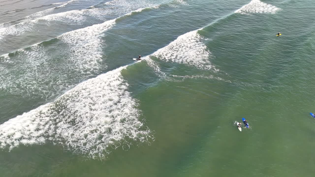 Aerial footage captures surfers paddling and riding gentle waves in bright daylight at West Sands Beach, St Andrews, Scotland. Smooth camera movement enhances the tranquil coastal atmosphere