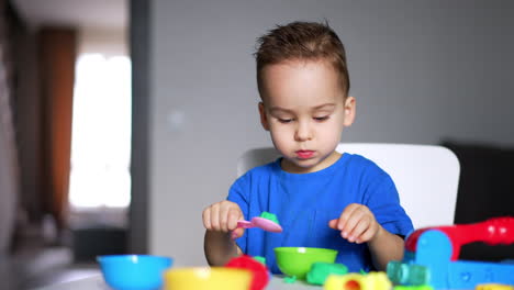 cute focused baby boy uses plastic cups and spoon in his game. calm caucasian toddler plays with plasticine. blurred backdrop.
