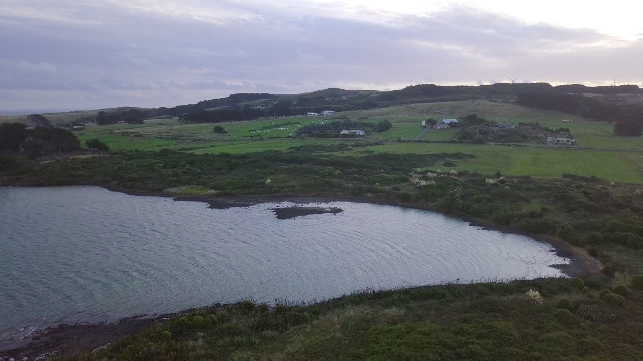 Aerial drone footage of farmland along the coast of New Zealand at sunset with windmills in the background
