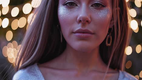 fashionable young woman wearing a glittering silver dress is posing in an outdoor setting with warm string lights creating a magical bokeh effect in the background