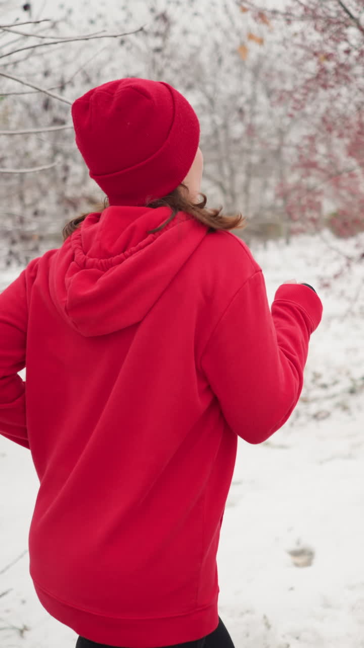 lady wearing red hoodie and beanie jogging along paved snow covered path outdoors during winter amid serene park scenery with barren trees and benches, hair swaying in motion conveying energy