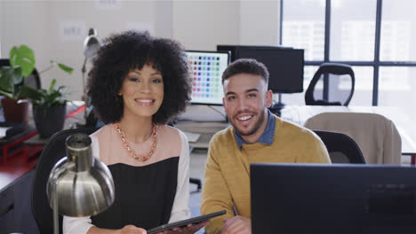 portrait of happy diverse male and female colleague at desk using tablet and computer, slow motion