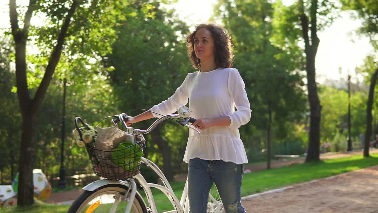 ⁇  셔츠 와 파란 청바지 를 입은 매력적 인 여자 가 도시 공원 의 골목 을  ⁇ 고 도시 자전거  ⁇ 바 를 들고 있다