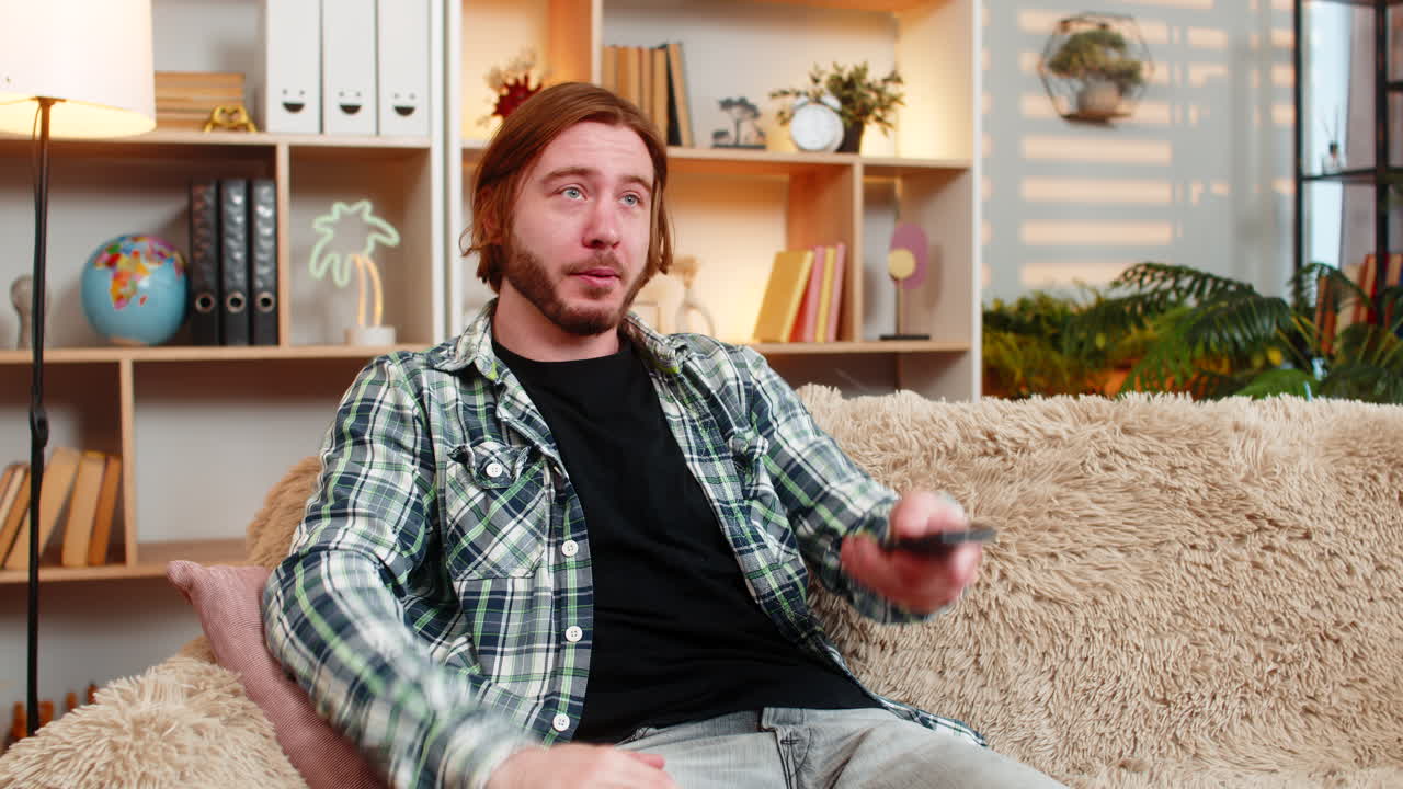 Giovane uomo sul divano di casa cambia i canali TV con il telecomando, poi reagisce a una scena TV emozionante, in casa.