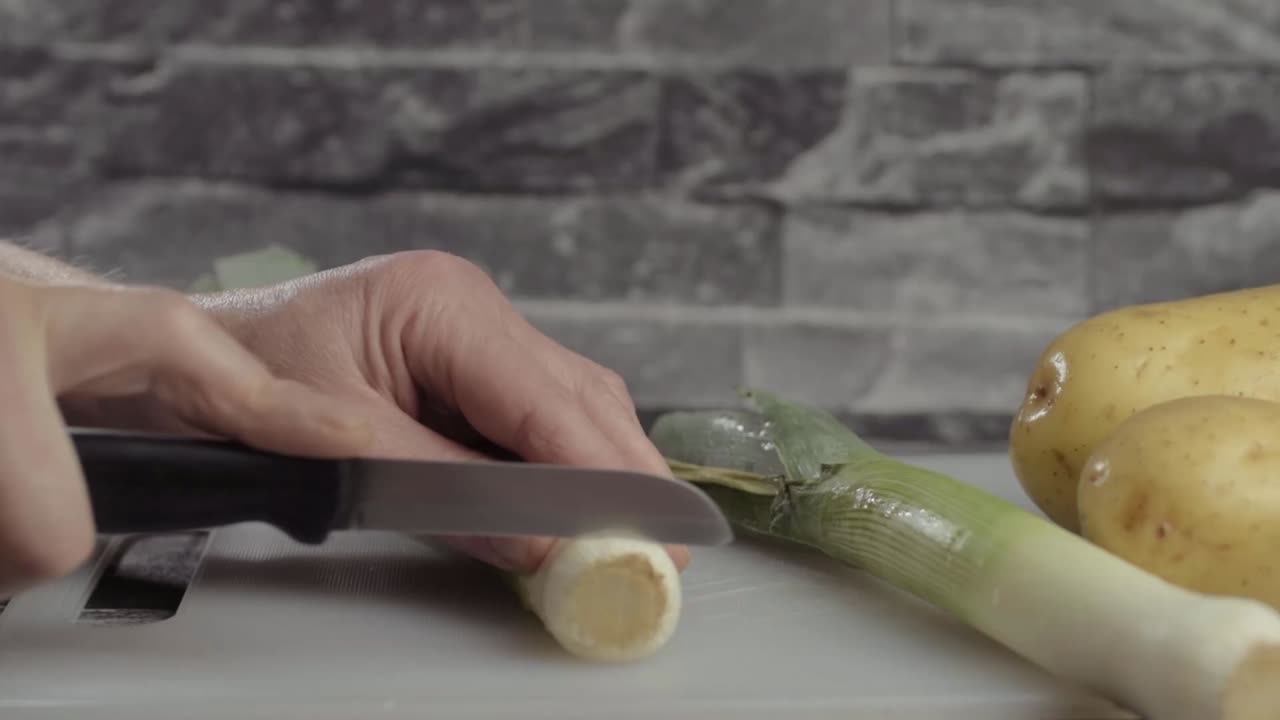 Hand slicing leek vegetable with potatoes in background medium shot