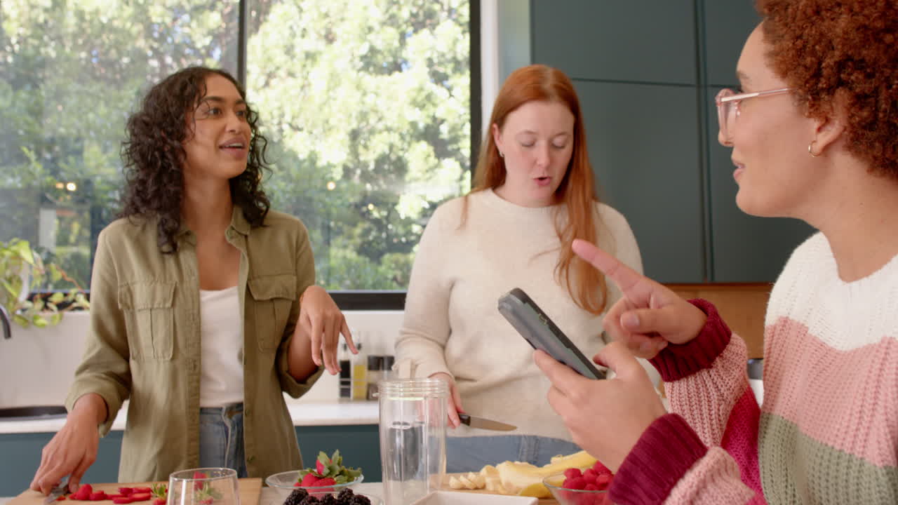 female friends hanging out in kitchen, talking and using smartphone, enjoying snacks