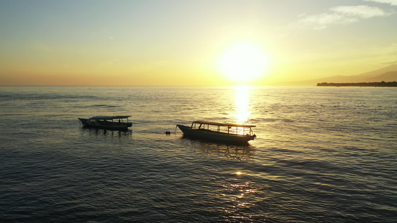 Fishing boats floating over calm surface of sea near shore of tropical island at sunset on a dusty sky with yellow sun over silhouette of mountains in Bali